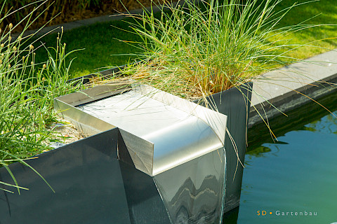 Individuell gefertige Wasserschütte aus Edelstahl in einem Metallhochbett mit Gräsern in einem Garten in Paderborn.